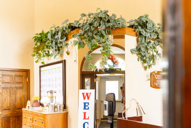 Interior lobby showing a welcome sign, leafy plants draped over an archway, wooden furniture, and a beverage station.