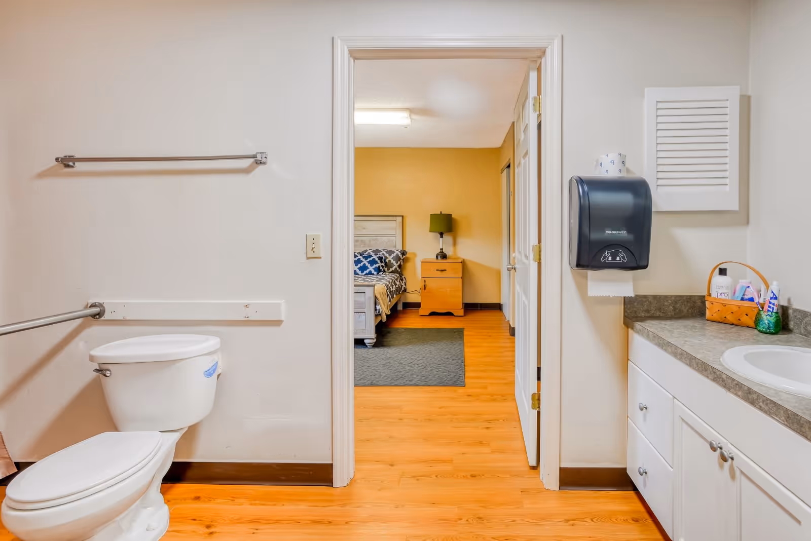 Bathroom with a toilet and sink countertop looking through an open doorway into a bedroom with a bed and nightstand.