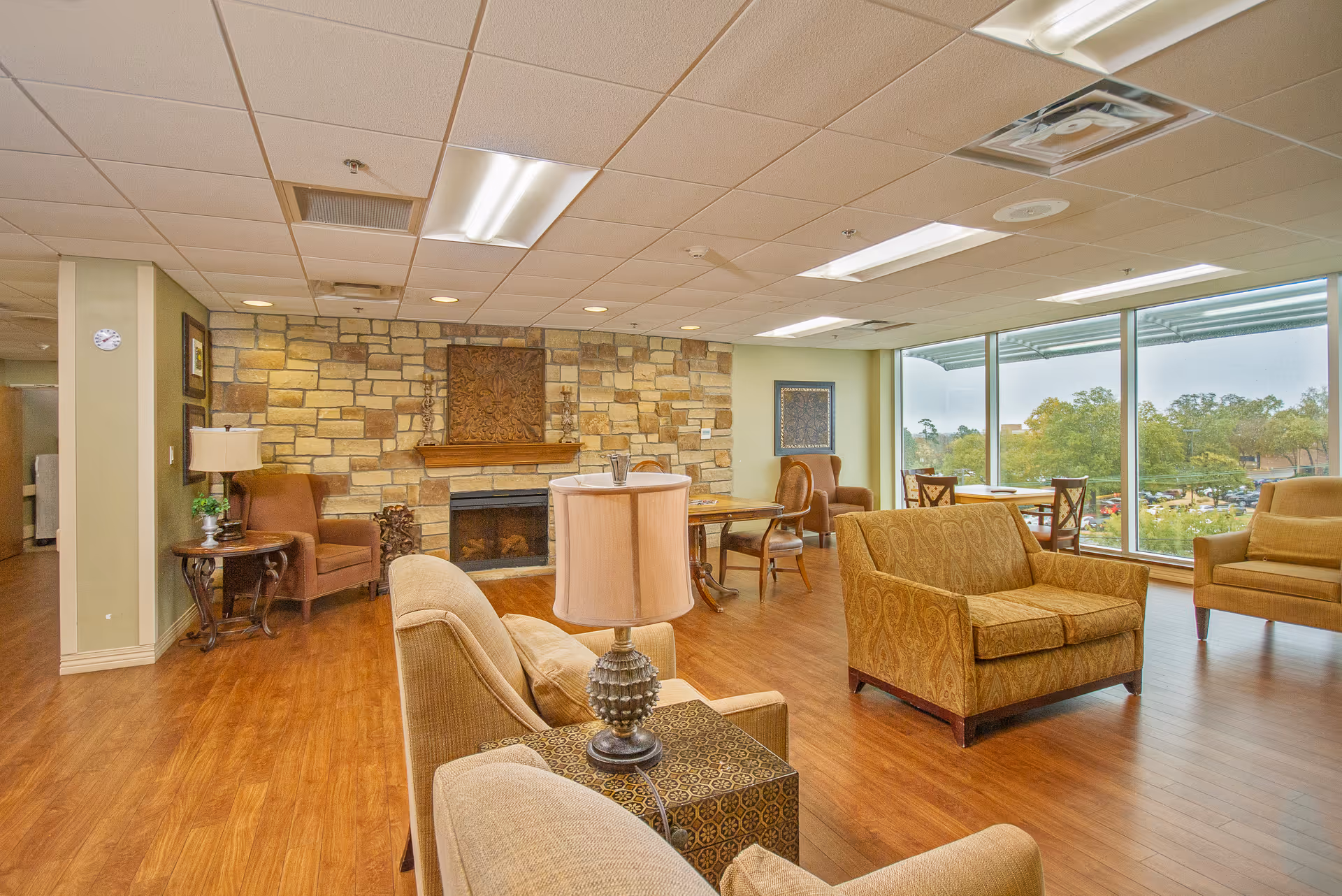 A spacious living room area with wooden flooring, beige and brown upholstered chairs and sofas, a stone fireplace with decorative art above it, and large windows offering a view of trees and a parking lot outside. The room is well-lit with ceiling lights and has a cozy, inviting atmosphere.