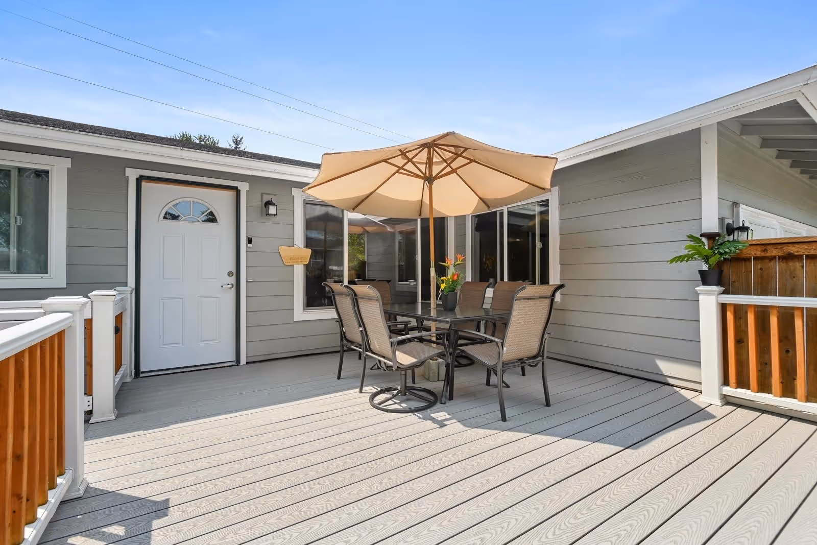 Sunlit outdoor deck with a glass table, six chairs and a large umbrella next to a gray-sided house entrance and railing.
