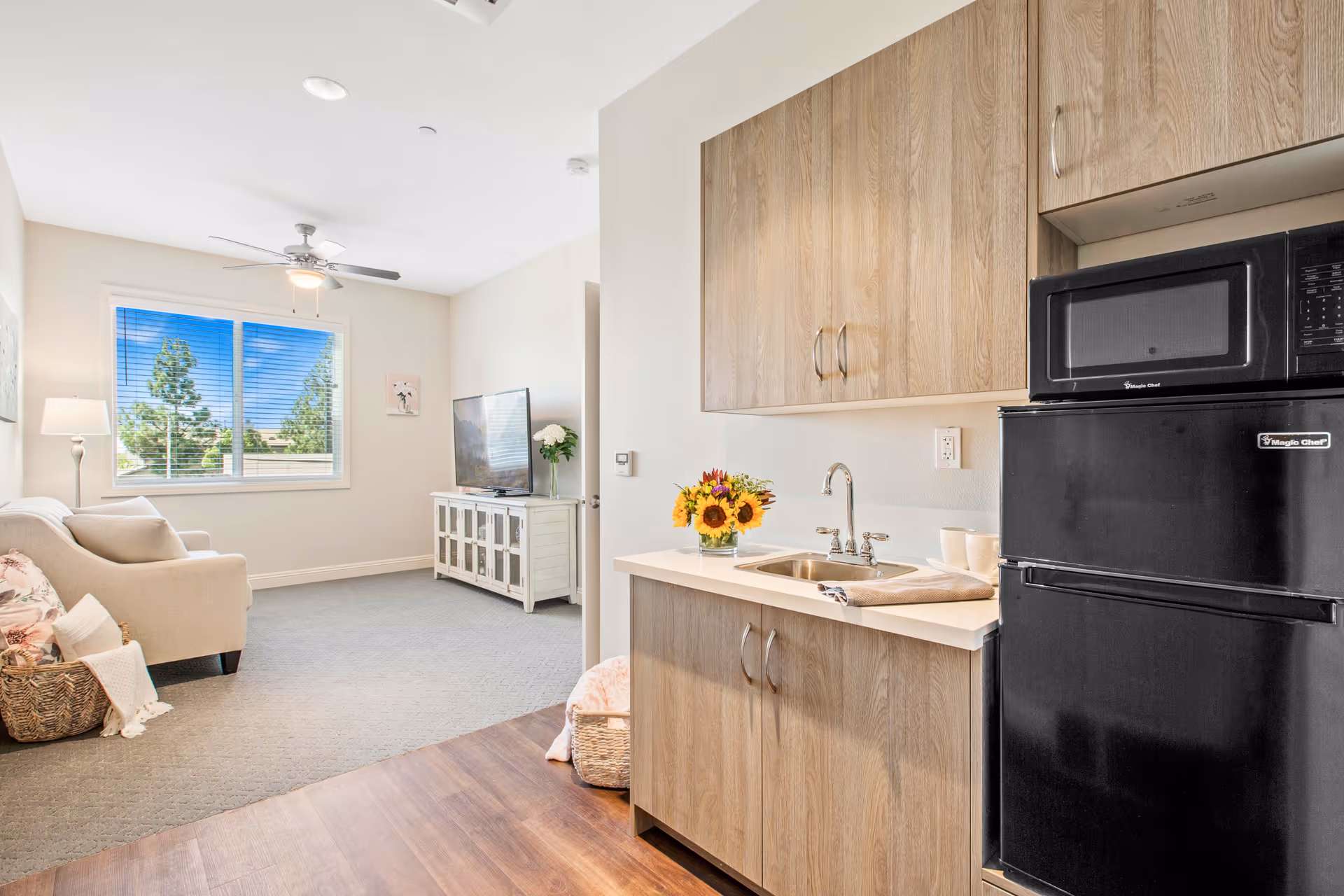 A bright and cozy living space featuring a small kitchenette with wooden cabinets, a sink, a black microwave, and a black mini refrigerator. The living area has a beige sofa with cushions and a basket with a blanket, a floor lamp, a window with blinds showing trees outside, a ceiling fan with light, and a white TV stand with a flat-screen TV and a vase of flowers.