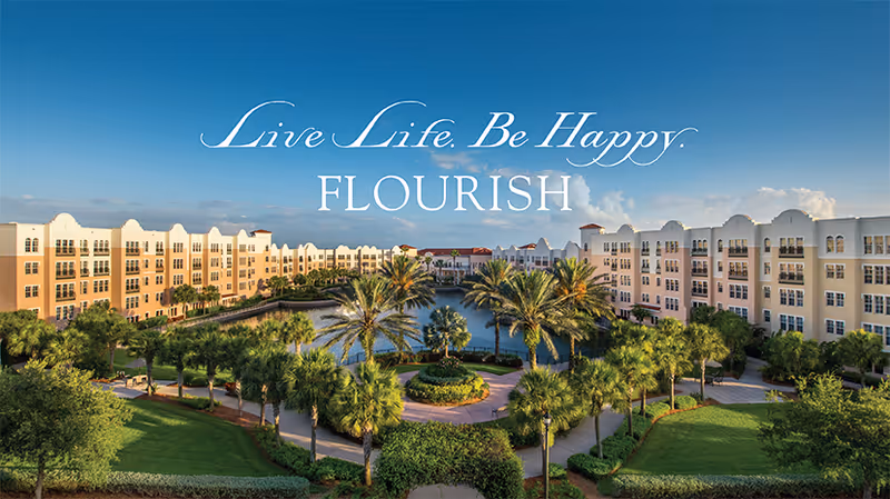 Aerial view of a senior living facility with two large beige buildings facing each other across a landscaped courtyard featuring palm trees, green lawns, and a central water feature under a clear blue sky.
