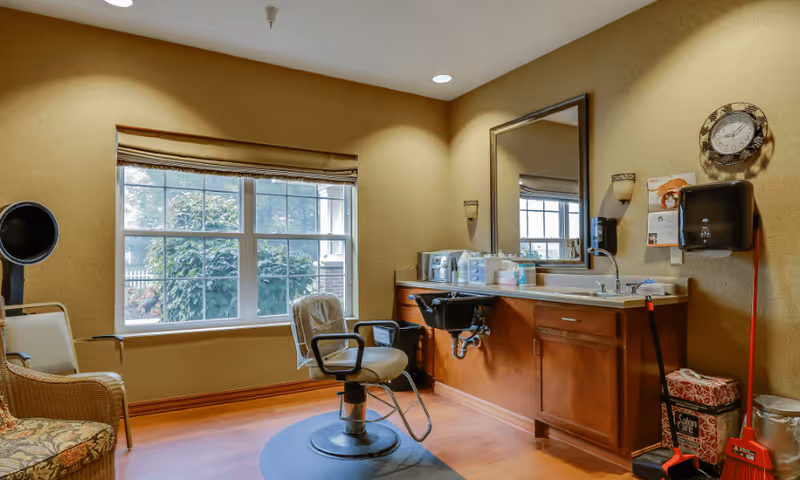 A small salon room with a styling chair in front of a sink and mirror. The room has a large window with a view of greenery outside, beige walls, wooden cabinetry, and various salon supplies. There is a clock on the wall, a paper towel dispenser, and cleaning tools in the corner.