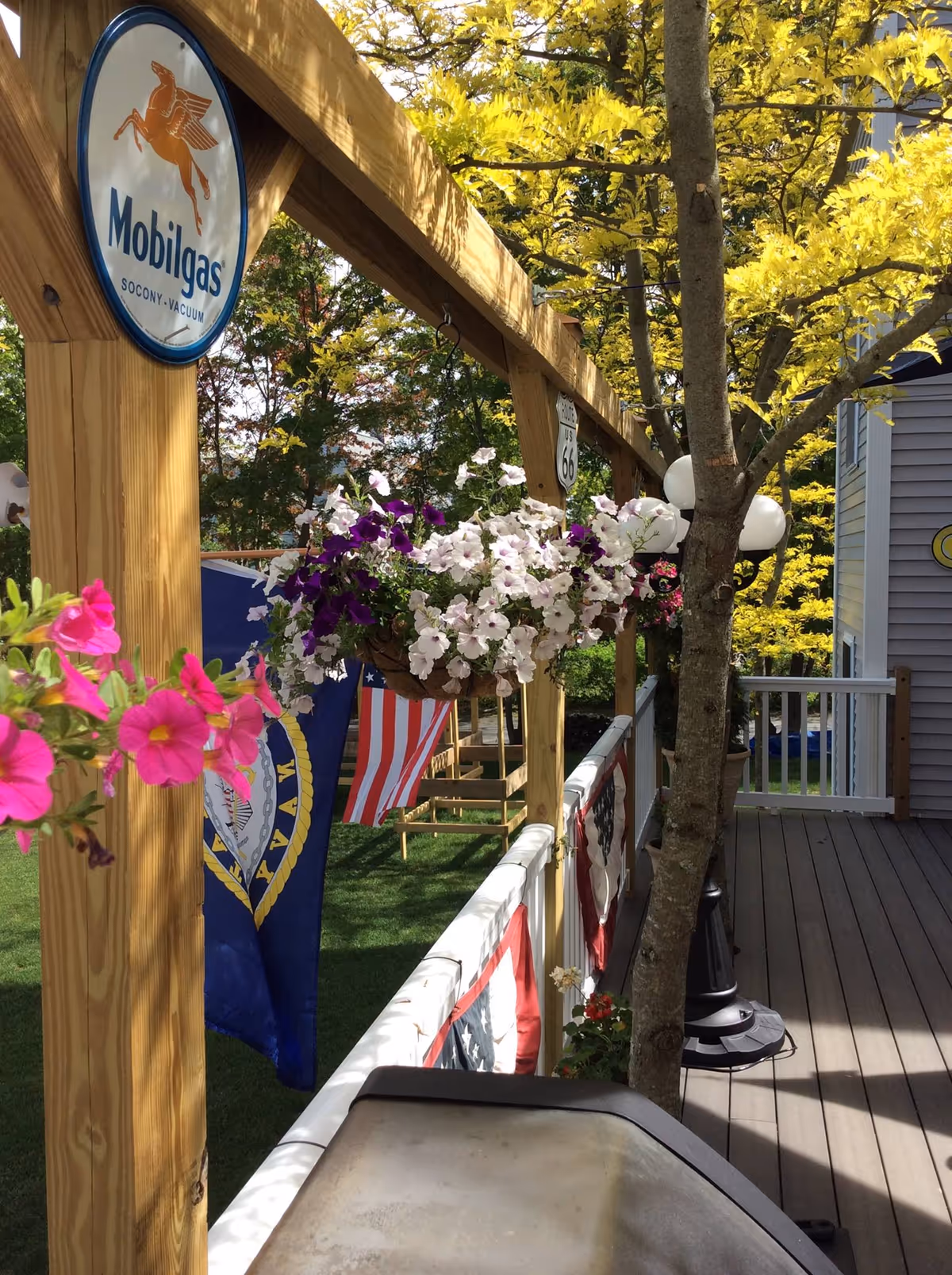A wooden deck railing decorated with hanging flower baskets containing white and purple flowers, and pink flowers in the foreground. There are American and Navy flags hanging from the railing. A tree grows through the deck near a black lamp post with white globe lights. A Mobilgas sign and a Route US 66 sign are attached to the wooden beams above the railing. Green grass and trees with yellow leaves are visible in the background.