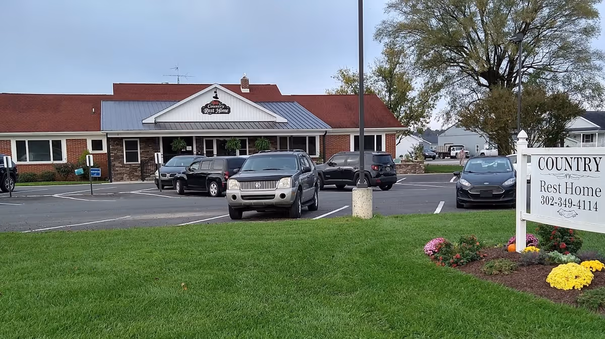 Exterior view of Country Rest Home building with a parking lot in front containing several parked cars. There is a green lawn with a flower bed and a white sign displaying the facility name and phone number.