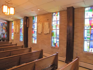 Interior chapel with wooden pews, stained glass windows set in brick walls, and overhead lighting.