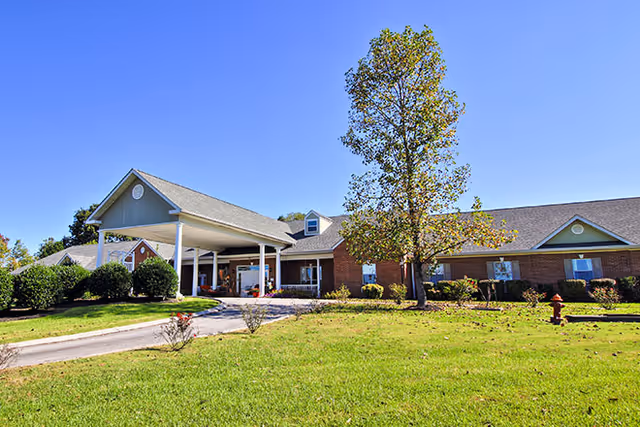 Exterior view of a single-story brick building with a covered entrance, surrounded by green grass, bushes, and a tree under a clear blue sky.