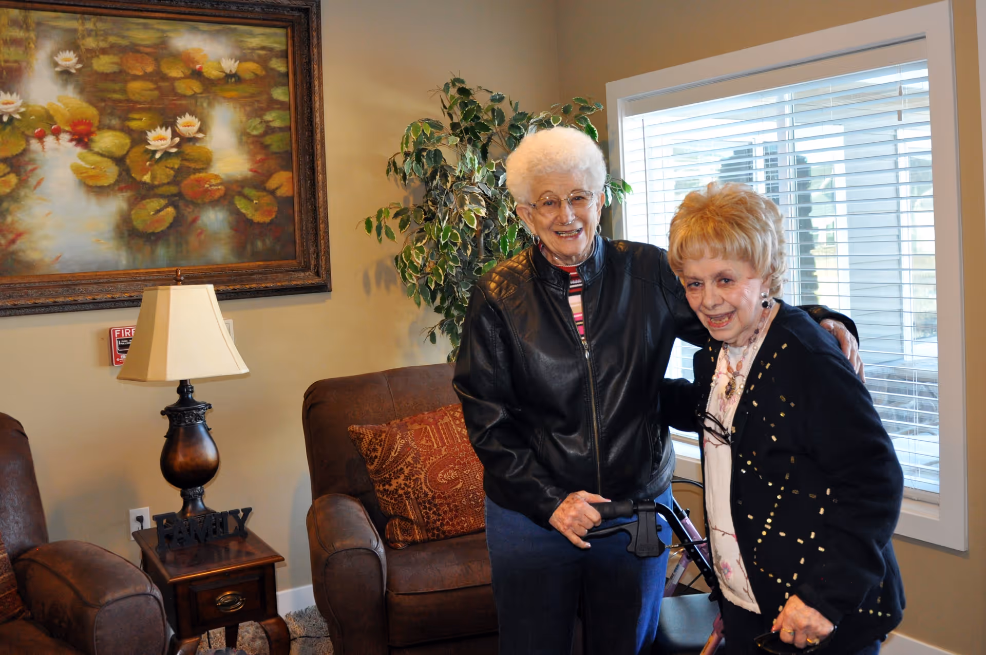 Two elderly women smiling and standing together in a comfortable living room with armchairs, a lamp, wall art, and a window.