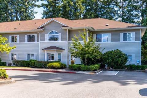 Exterior view of a two-story residential building with beige siding and a brown roof, surrounded by green bushes and trees, with a parking area in front.