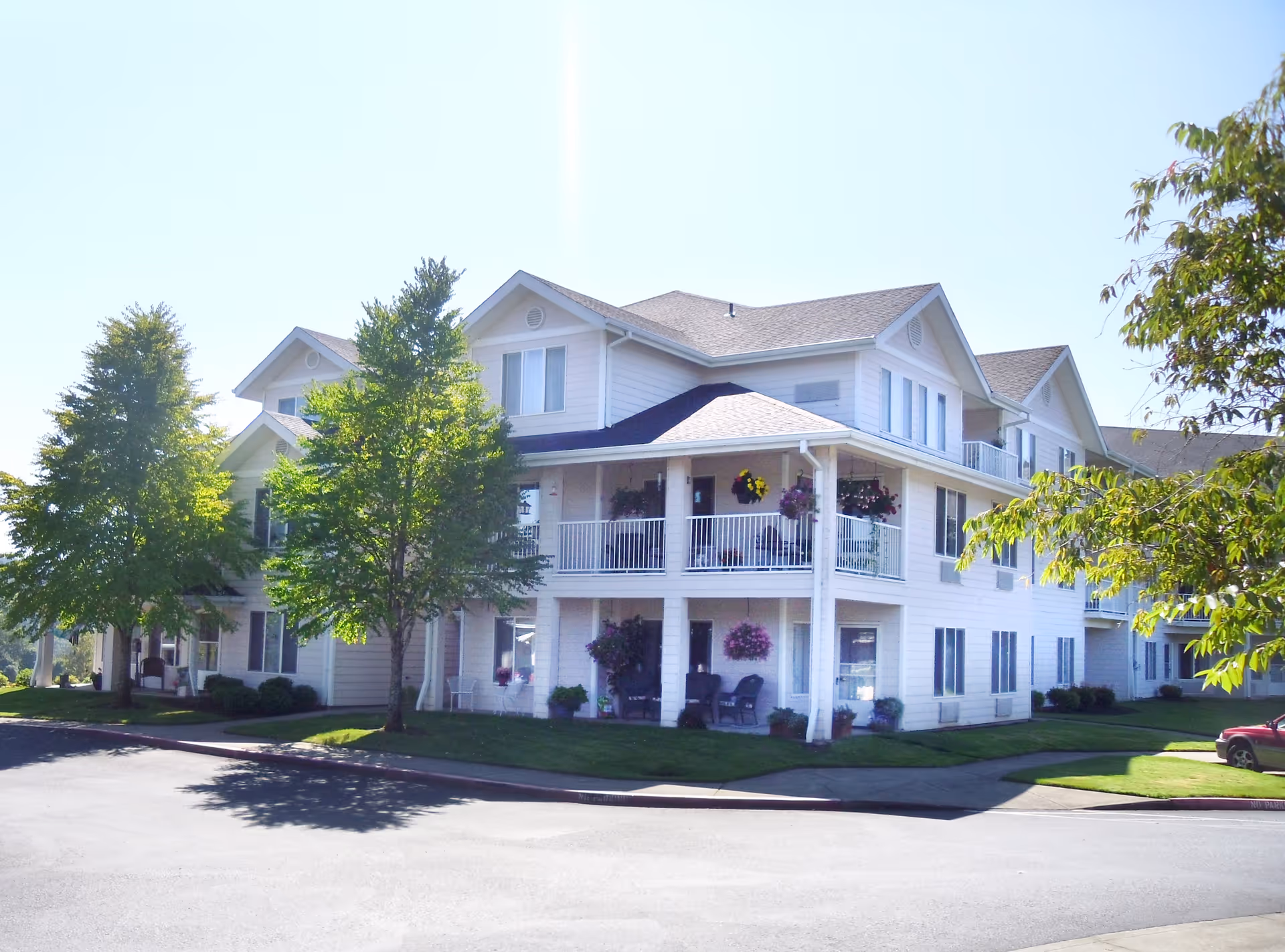 Three-story white residential building with covered balconies, hanging flower baskets, and landscaped trees.