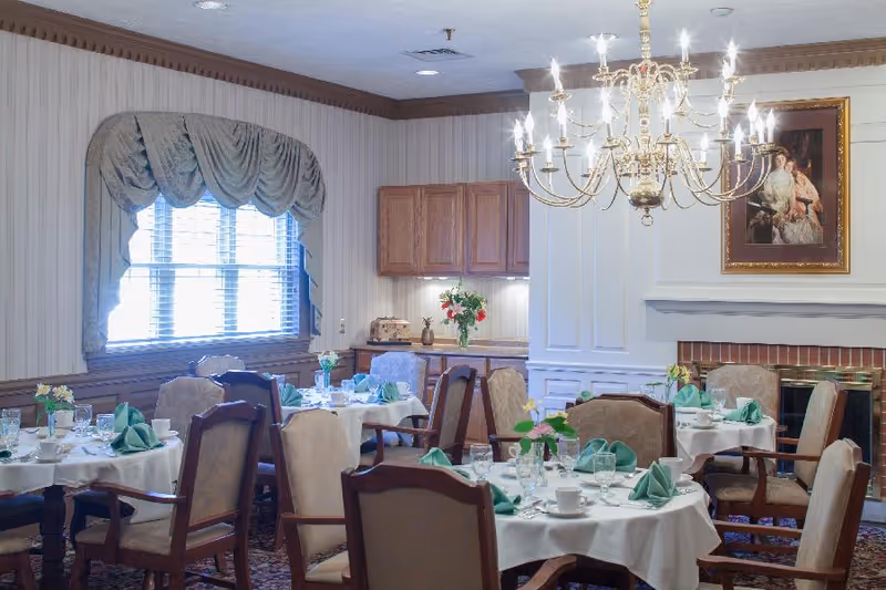 A dining room in a senior living community with round tables covered in white tablecloths, set with plates, cups, glasses, and green folded napkins. The room features upholstered chairs, a large chandelier, a window with draped curtains, wooden cabinetry, and a fireplace with a framed portrait above it.