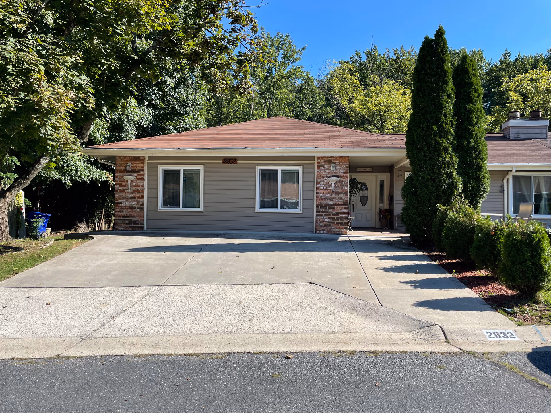 Single-story assisted living house front with a wide concrete driveway, two front windows, brick accents and tall shrubs by the entrance.