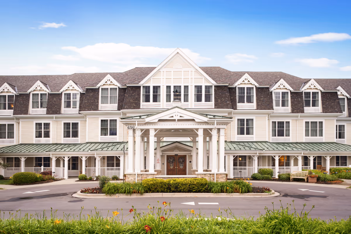 Front exterior view of a large, multi-story senior living facility building with a covered entrance, multiple windows, and a landscaped driveway with flowers and greenery.