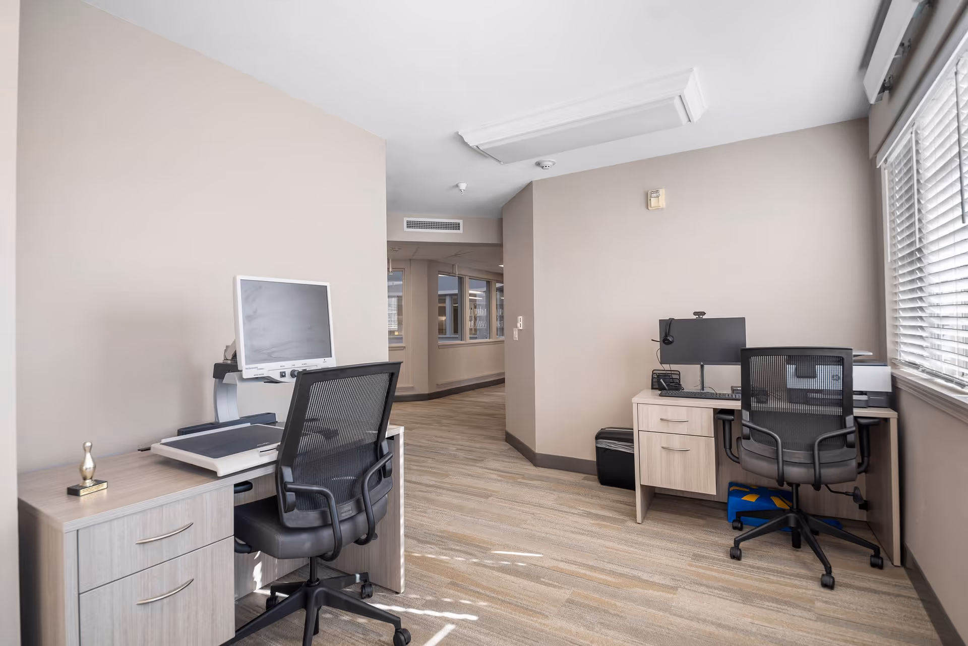 A small office area with two workstations, each equipped with a computer monitor and office chair. The room has beige walls, a window with blinds on the right side, and light wood flooring. The space is well-lit with natural light coming through the window.
