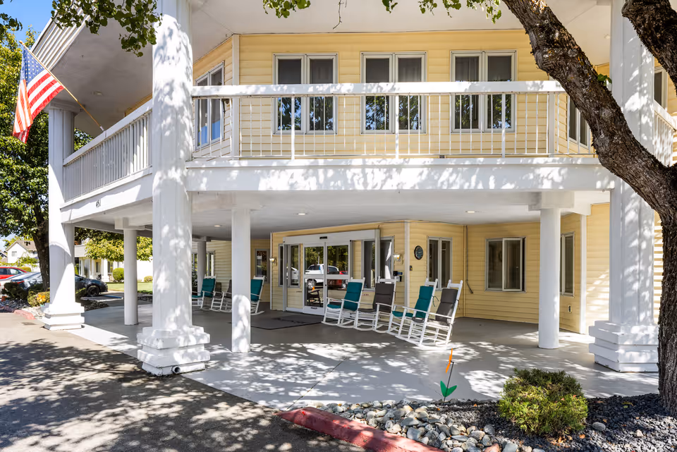 Entrance of a senior living community building with a covered porch supported by white columns. Several rocking chairs are arranged on the porch near the glass entrance doors. The building exterior is light yellow with white trim. An American flag is mounted on one of the columns. Trees and landscaping are visible around the entrance area.