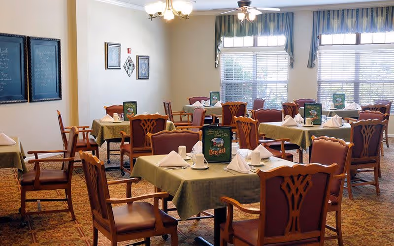 A dining room with multiple tables covered in green tablecloths, each set with white napkins, cups, and menus. The room has wooden chairs with cushioned seats and backs, large windows with striped valances, and framed artwork on the walls.