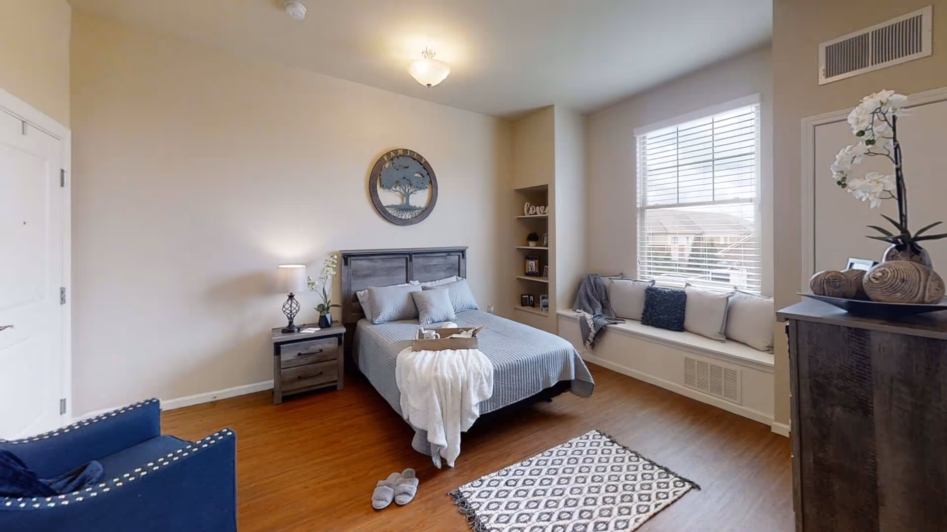 Bright furnished bedroom with a bed, nightstand, window seat, armchair, and dresser.