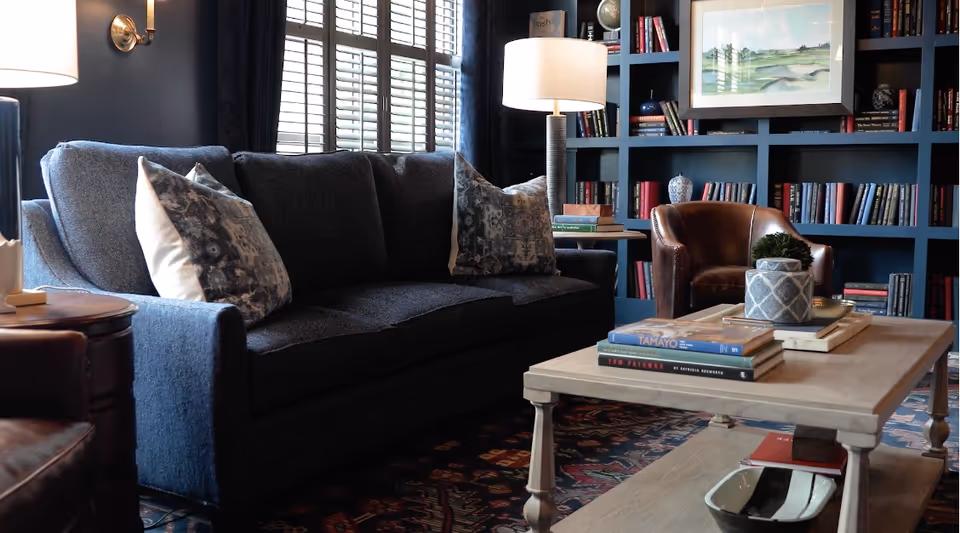 Cozy living room with a dark upholstered sofa, coffee table stacked with books and decor, leather chairs, lamps, and a built-in bookshelf wall.