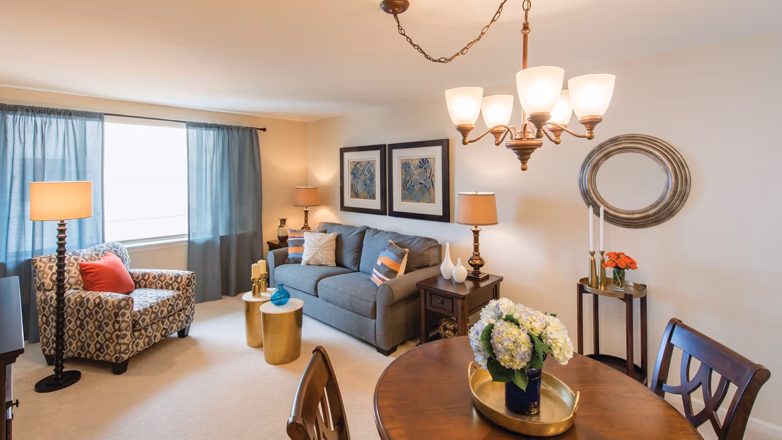 A cozy living room with a patterned armchair with a red pillow, a gray sofa with decorative pillows, two side tables with lamps, two framed artworks on the wall, a round mirror, a small table with candles and flowers, and a wooden dining table with chairs and a flower arrangement under a chandelier.