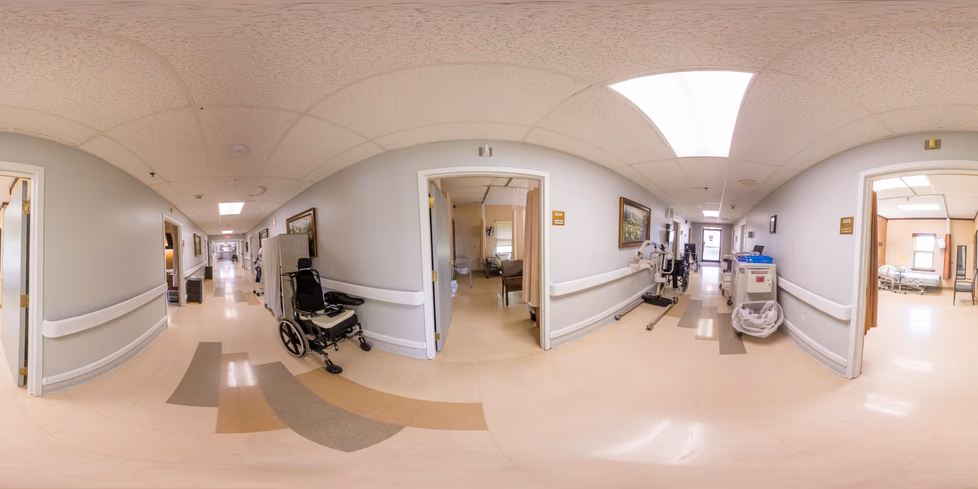 A wide-angle view of a clean, well-lit nursing home hallway with beige floors and light gray walls. Several open doors reveal patient rooms with hospital beds and medical equipment. Wheelchairs and medical carts are positioned along the hallway walls. Fluorescent ceiling lights illuminate the corridor, and handrails run along both sides of the hallway.