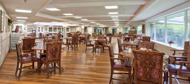 Bright, spacious dining room with multiple round tables and upholstered wooden chairs on a wood floor and large windows along one side.