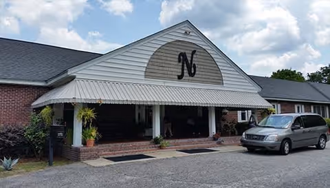 Front exterior view of Northwoods Senior Living building with a large awning featuring a decorative 'N' above the entrance. A minivan is parked in front on a paved driveway under a partly cloudy sky.