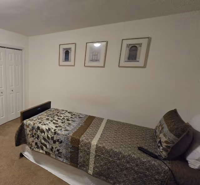 Small bedroom with a single bed against a wall, patterned bedspread, three framed pictures above and a closed closet.