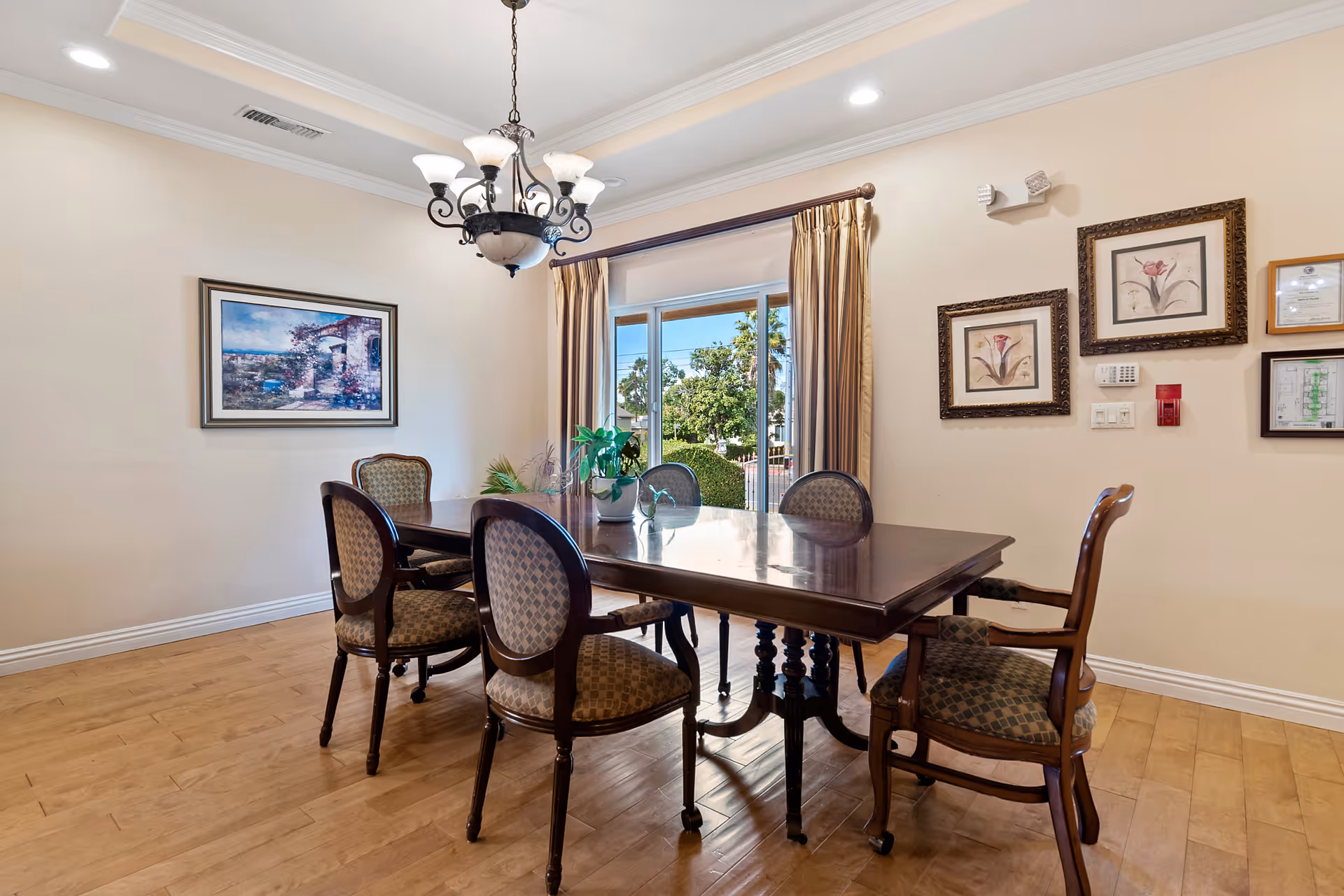 A dining room with a wooden table and six upholstered chairs. A chandelier hangs above the table. There are framed pictures on the beige walls and a large window with curtains showing greenery outside.