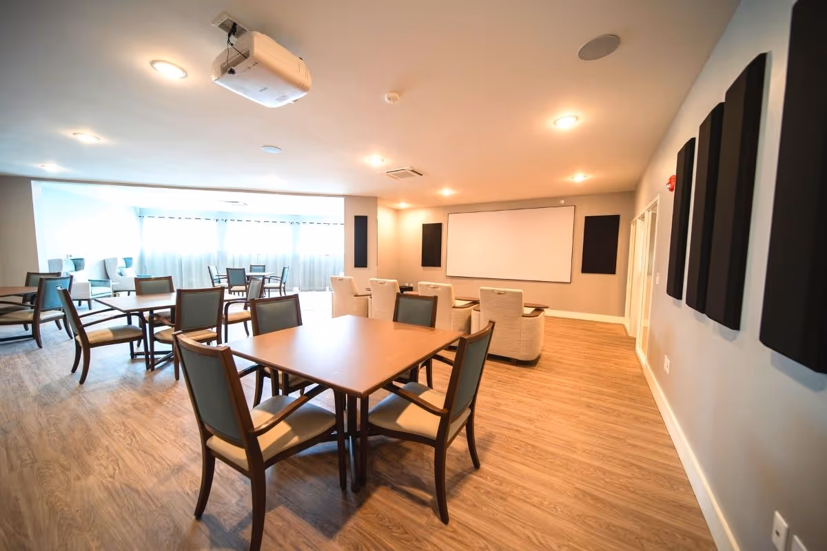 A spacious, well-lit room with multiple wooden tables and chairs arranged for group seating. The room features a projector mounted on the ceiling, a large white projection screen on the far wall, and several cushioned armchairs facing the screen. The floor is covered with wood-patterned flooring, and the walls have sound-absorbing panels. Large windows with sheer curtains allow natural light to brighten the space.