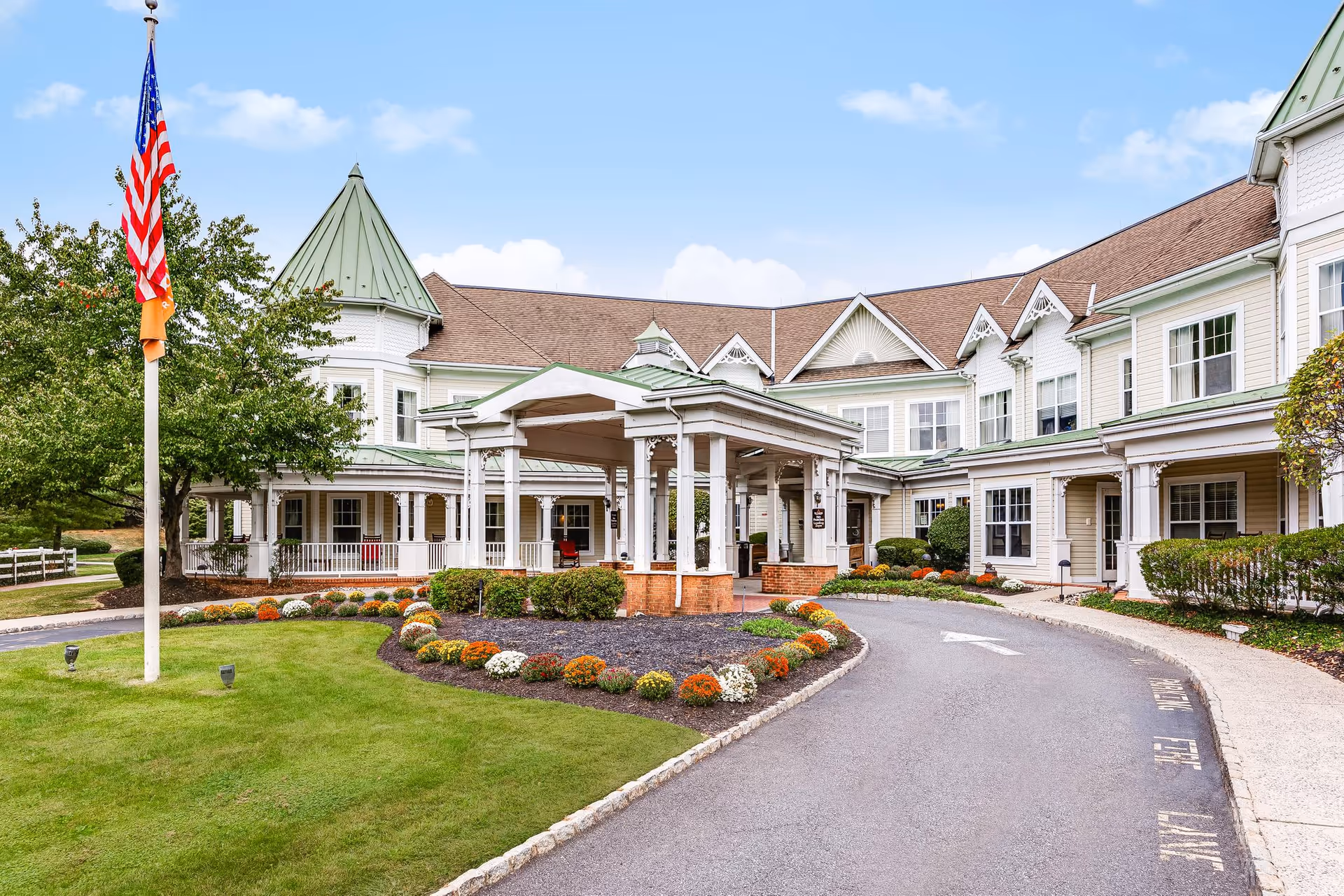 Exterior view of Sunrise of Westfield senior living facility showing a large building with white siding, green roofs, and a covered entrance. There is a circular driveway with landscaped flower beds and an American flag on a flagpole on the left side.