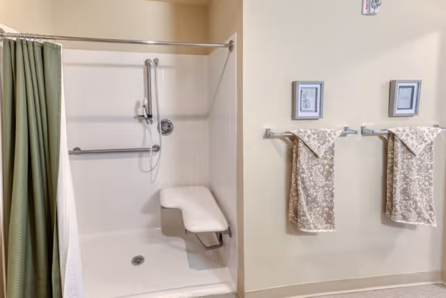 Accessible shower stall with a fold-down bench, grab bars and a green curtain next to two patterned towels on a beige wall.