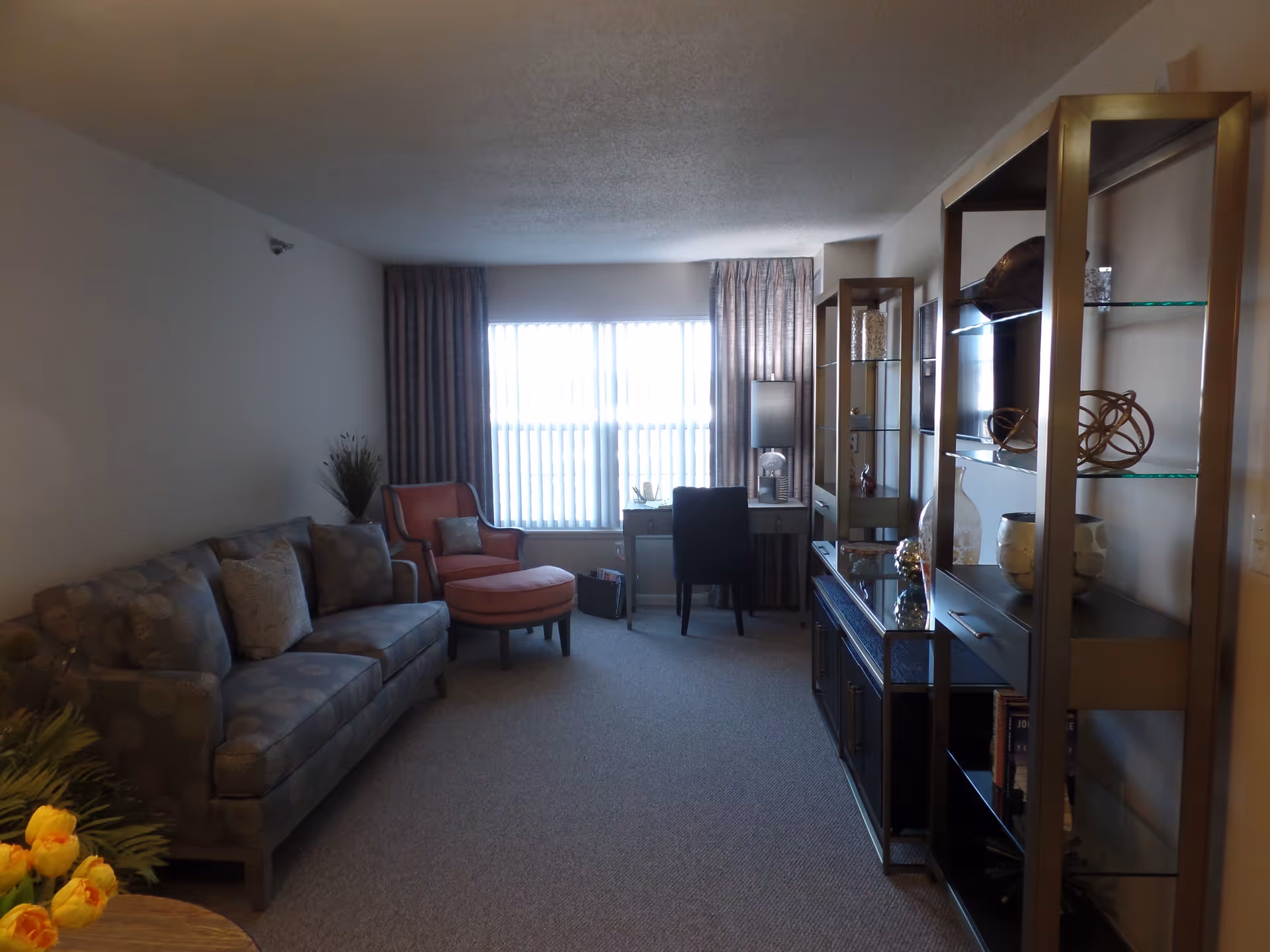 A living room with a patterned sofa and cushions on the left, an orange armchair with an ottoman near a window with vertical blinds and curtains. A desk with a black chair and a lamp is positioned in front of the window. On the right side, there is a shelving unit with decorative items and a TV. The room has carpeted flooring and neutral-colored walls.