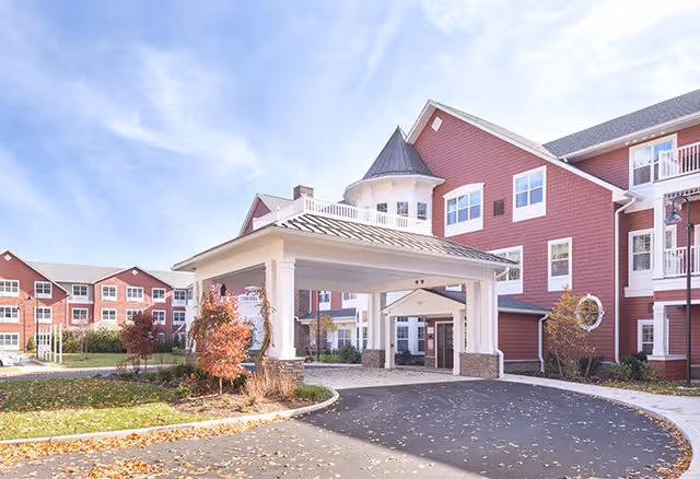 Exterior view of a senior living facility with red siding and white trim. The building has multiple windows and balconies, with a covered entrance driveway. There are trees with autumn foliage and a clear sky above.