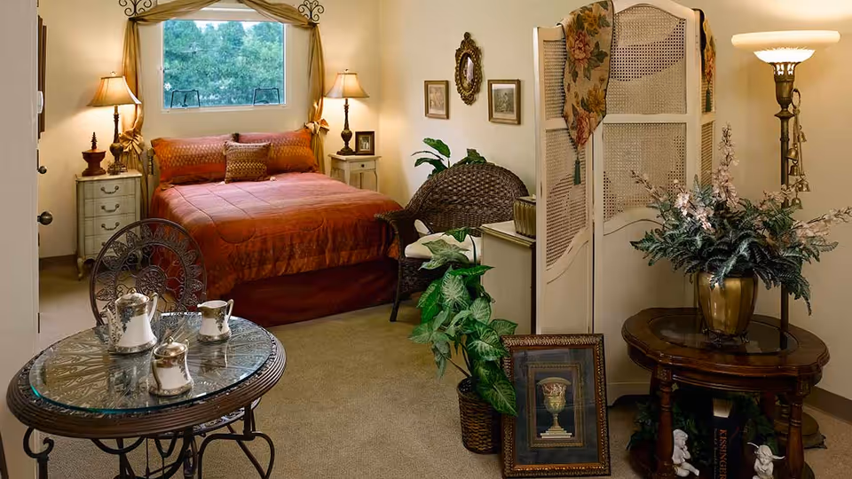 A cozy bedroom with a bed covered in a rust-colored quilt and matching pillows, flanked by two nightstands each with a lamp. A small round glass-top table with a tea set is in the foreground. The room also features a wicker chair, a decorative folding screen with a floral cloth draped over it, framed artwork on the walls, and potted plants. A window above the bed shows greenery outside.
