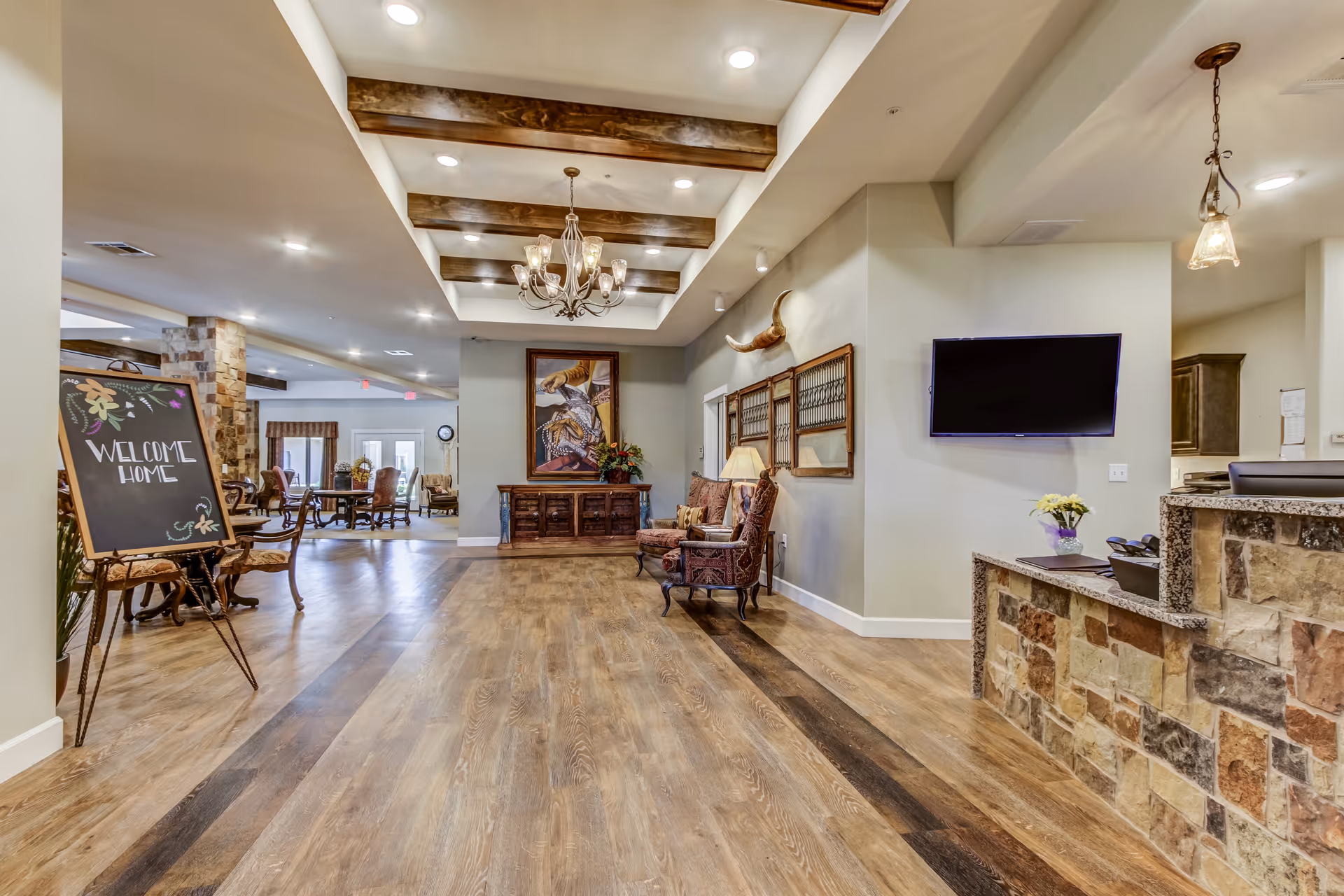 Interior view of a senior living facility lobby with wooden flooring, a stone reception desk, a wall-mounted TV, two upholstered chairs with a lamp, a large painting, decorative wall hangings, and a chandelier with wooden ceiling beams. A chalkboard sign reads 'Welcome Home' near a seating area with tables and chairs in the background.