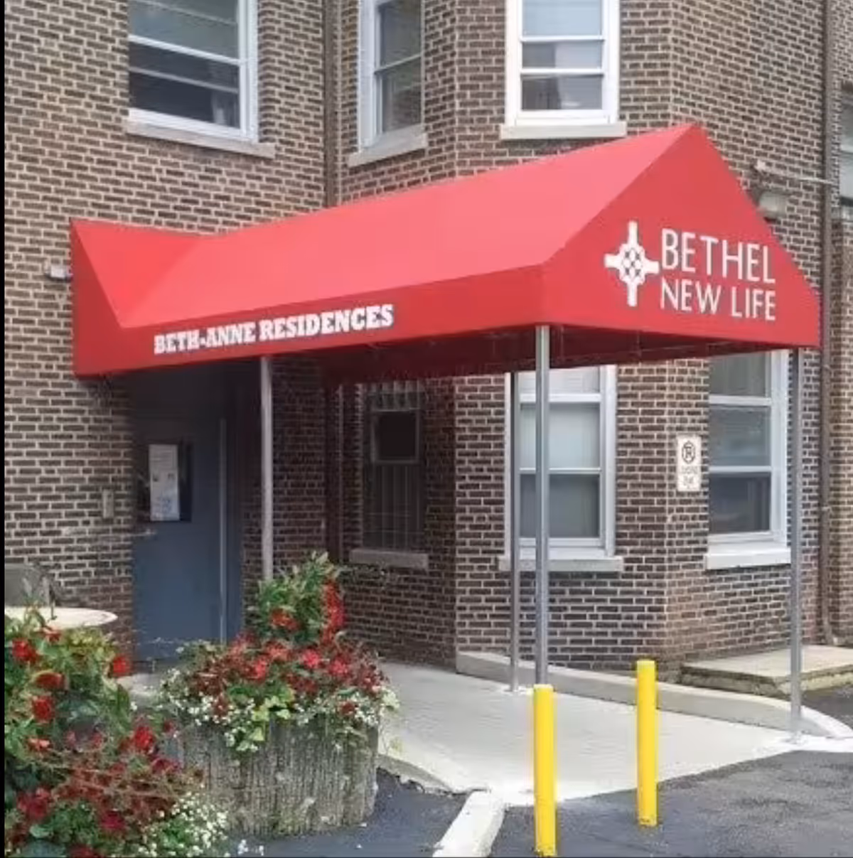 Entrance to a brick building with a red canopy that reads 'BETHEL-ANNE RESIDENCES' on the side and 'BETHEL NEW LIFE' on the front. There are two yellow bollards in front of the entrance and a planter with red and green plants nearby.