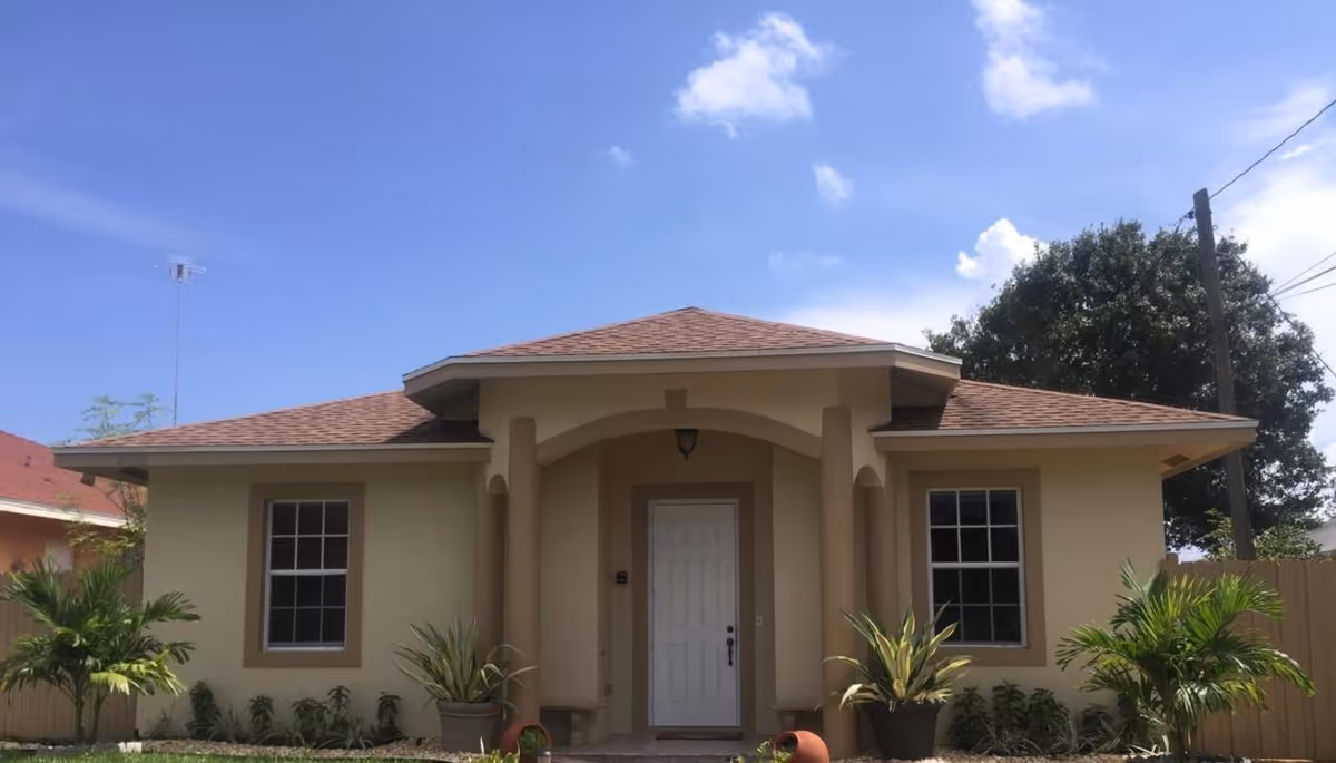 Front exterior of a single-story beige house with a centered white door, flanking windows, columns, and potted plants under a blue sky.