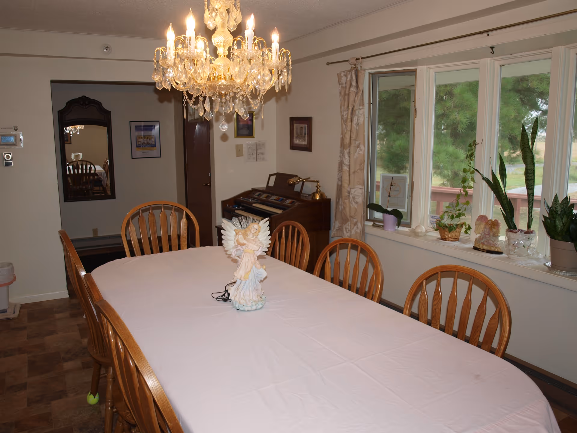 A dining room with a long table covered with a white tablecloth and surrounded by wooden chairs. A decorative angel figurine is placed in the center of the table. Above the table hangs a crystal chandelier with lit bulbs. To the right, there is a large window with several potted plants on the windowsill and beige curtains. In the background, there is a wooden organ against the wall and a mirror reflecting part of the room.