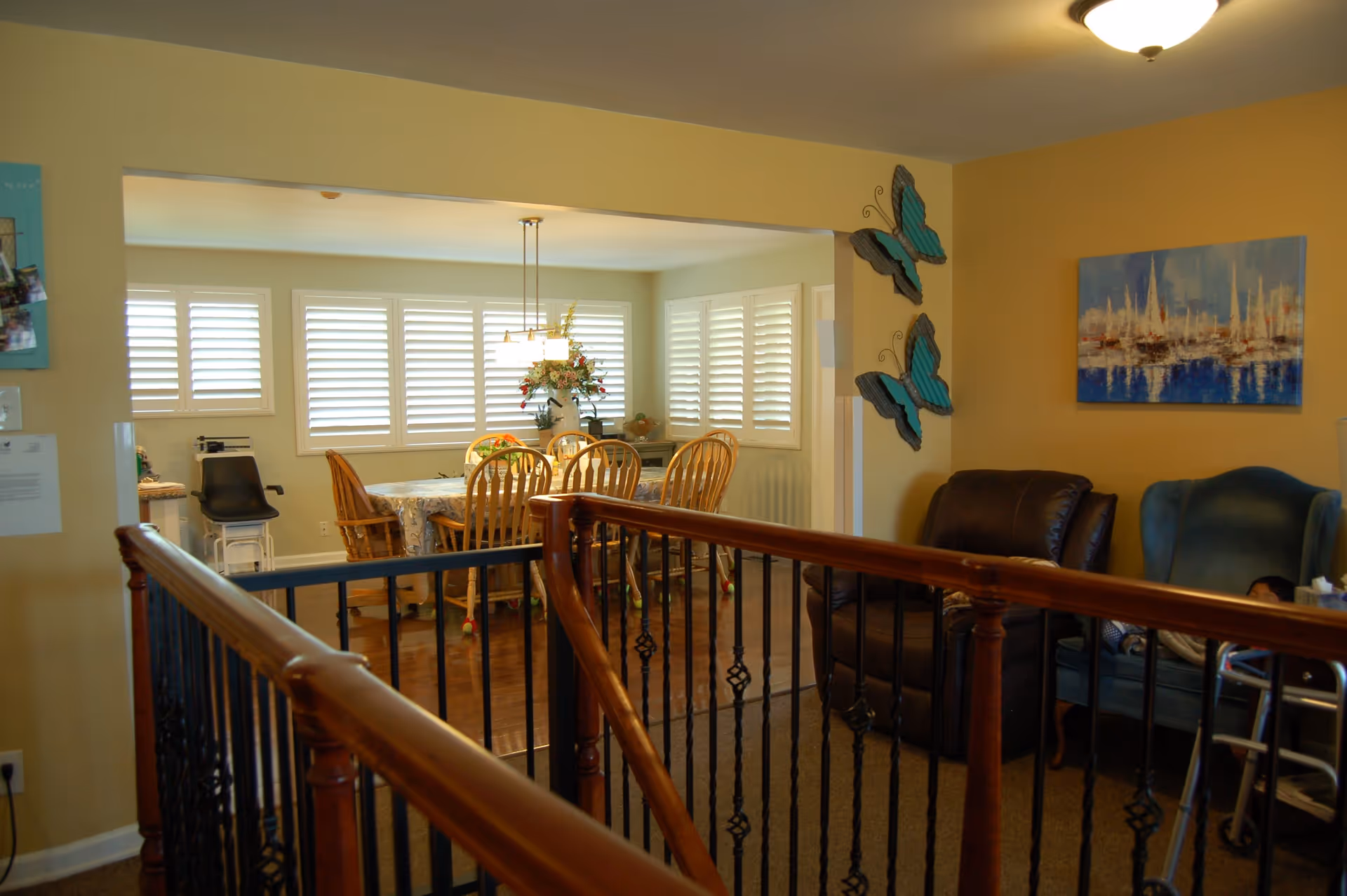 Interior view of a senior living facility showing a dining area with a table and chairs near windows with white shutters, and a sitting area with a leather chair and a blue armchair. There are two decorative blue butterflies on the wall and a painting of sailboats. A wooden railing with black metal spindles is in the foreground.