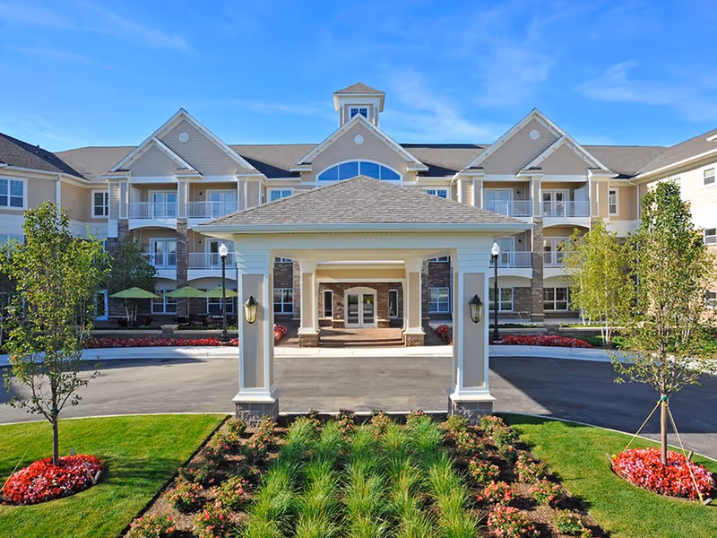 Front exterior view of a large senior living facility building with a covered entrance, landscaped garden with flowers and greenery, and a clear blue sky.