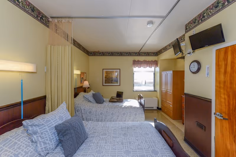 A well-lit bedroom in a senior living facility with two beds covered in gray patterned bedding and multiple pillows. There is a curtain divider between the beds, a window with blinds and a valance, a wooden wardrobe, a wall clock, two mounted flat-screen TVs, and a framed picture on the wall. The room has light yellow walls with a floral border near the ceiling.
