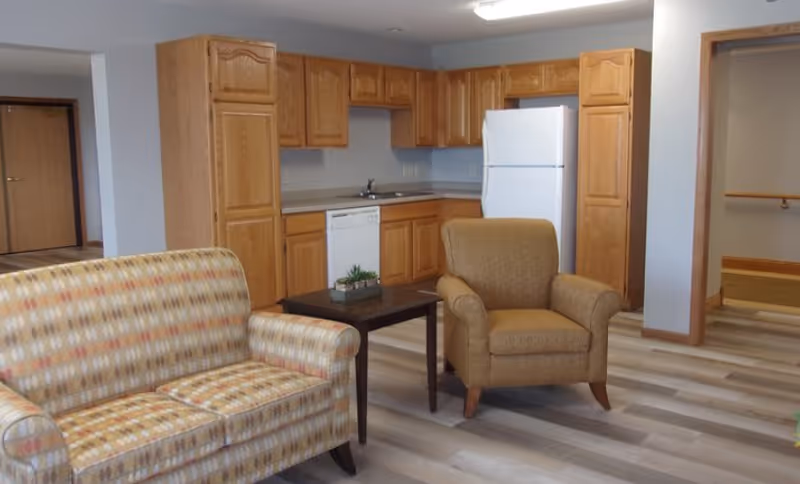 Open-plan living area with a patterned sofa, armchair and coffee table in front of a kitchenette with wooden cabinets and a refrigerator.