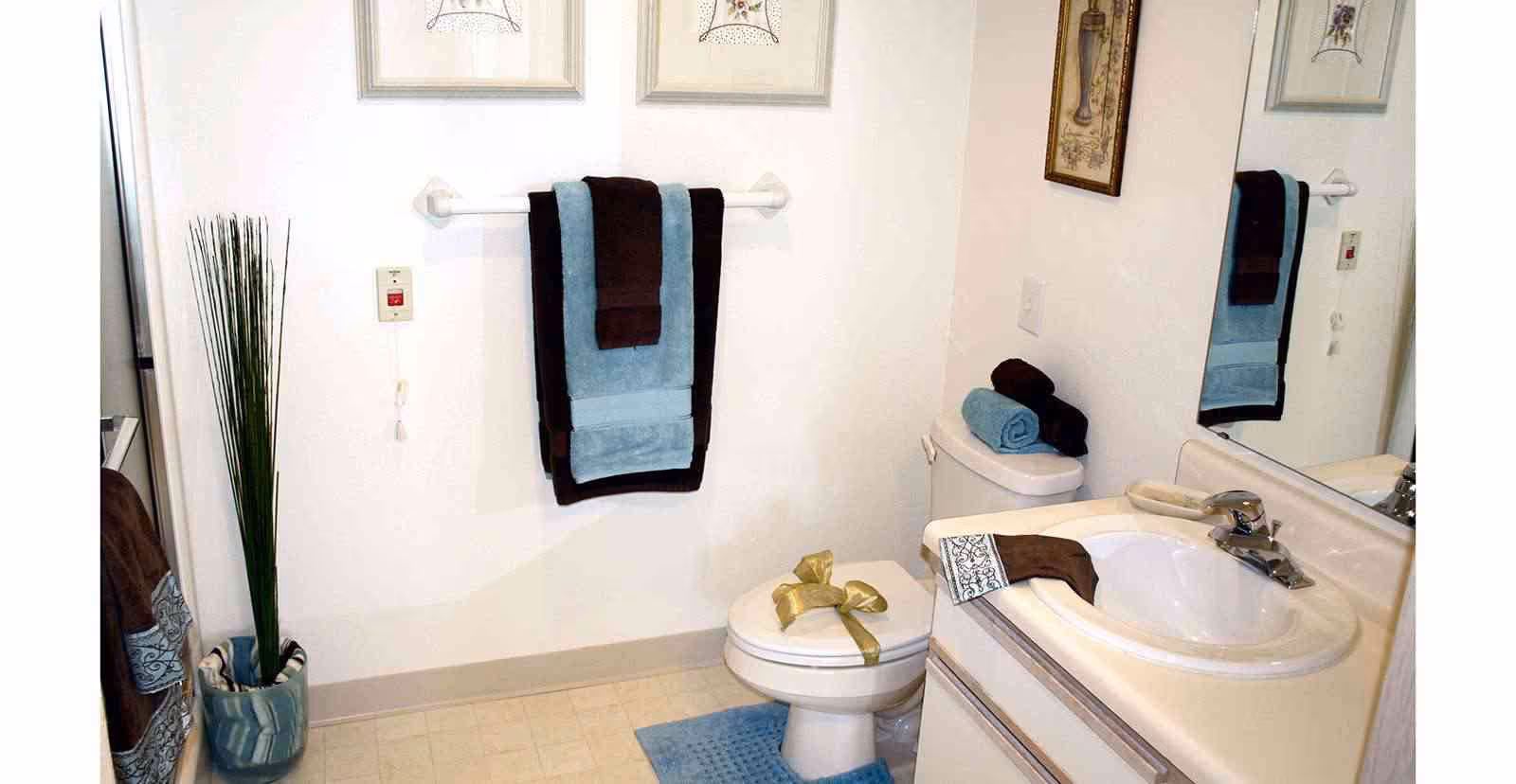 A clean bathroom with a white toilet decorated with a gold ribbon, a white sink with a silver faucet, and a mirror above the sink. There are neatly folded blue and brown towels on the towel rack and on the toilet tank. A blue bath mat is placed in front of the toilet, and a tall green plant in a blue pot is positioned near the wall. The walls are white with framed artwork hanging above the towel rack.