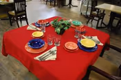 A dining table covered with a red tablecloth set for four people with colorful plates, glasses, and napkins, with a small green plant centerpiece. The background shows additional dining tables and chairs in a spacious room.