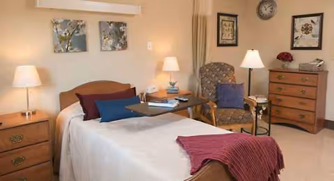 A furnished bedroom in a senior living facility with a bed, dresser, and tables.