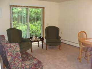 A living room with a floral patterned sofa, two green upholstered armchairs, a small wooden coffee table, and a round wooden dining table with chairs. There is a large window with vertical blinds showing greenery outside.