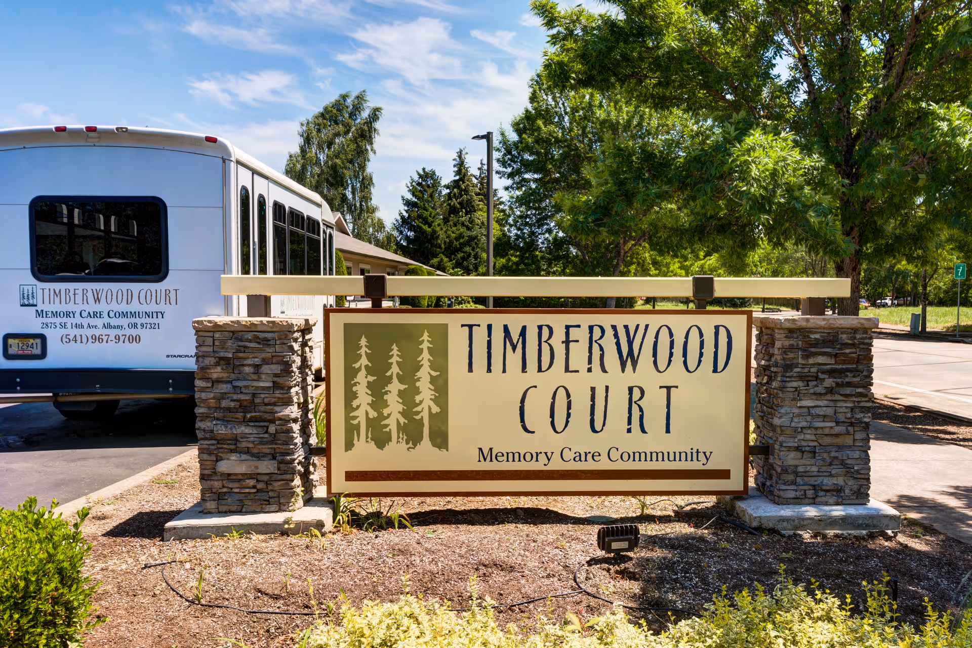 Entrance sign reading 'Timberwood Court Memory Care Community' between stone pillars with the facility shuttle and trees in the background.