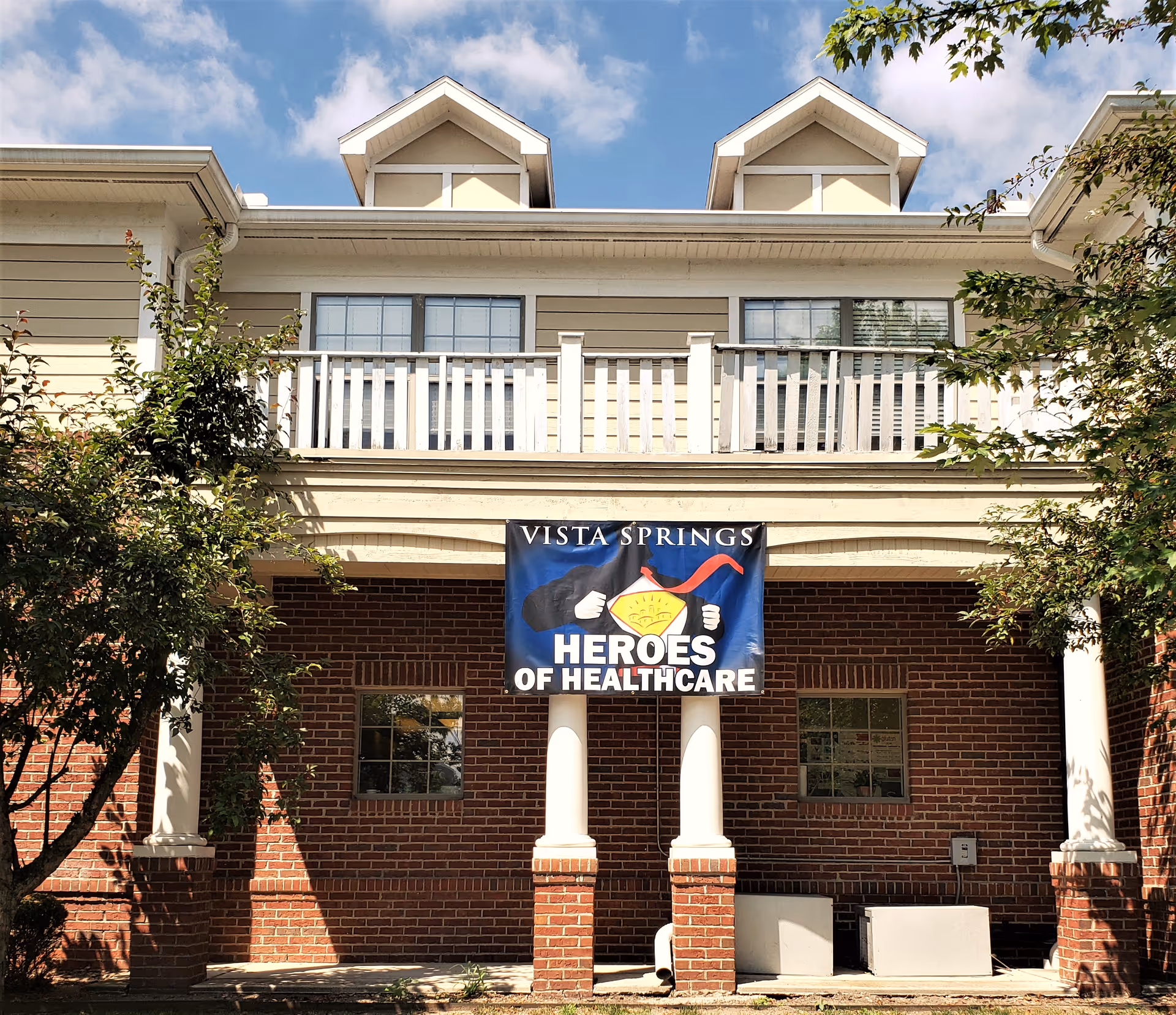 Exterior view of a brick and beige building with white columns and a balcony. A banner hanging between the columns reads 'Vista Springs Heroes of Healthcare'. Trees partially frame the building under a blue sky with some clouds.