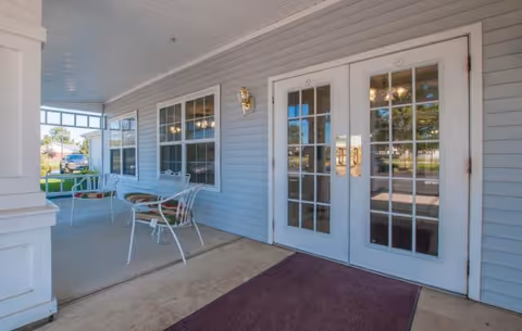 Covered outdoor patio area with white metal chairs and tables, light blue siding walls, large windows, and double glass doors with gold handles. A maroon doormat is placed in front of the doors.