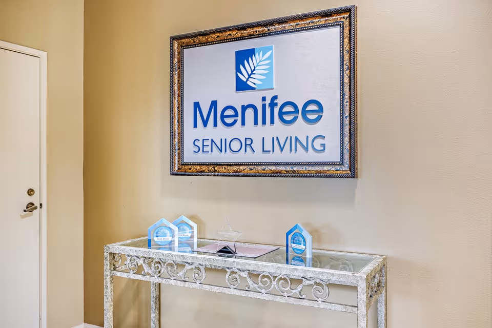 Framed "Menifee Senior Living" sign on a beige wall above a decorative glass-top console table with awards.